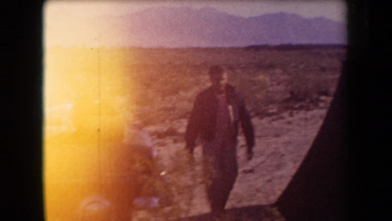 Adult man in a jacket crosses an arid Arizona landscape, partly obscured by strong flare and dark frame edge in the 8mm scan.