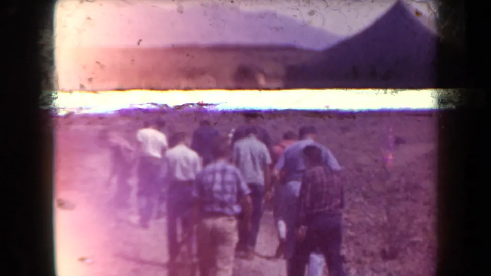 Group of adult men in 1958 Arizona walk away across rocky desert terrain, with strong exposure flare and hills in the distance.