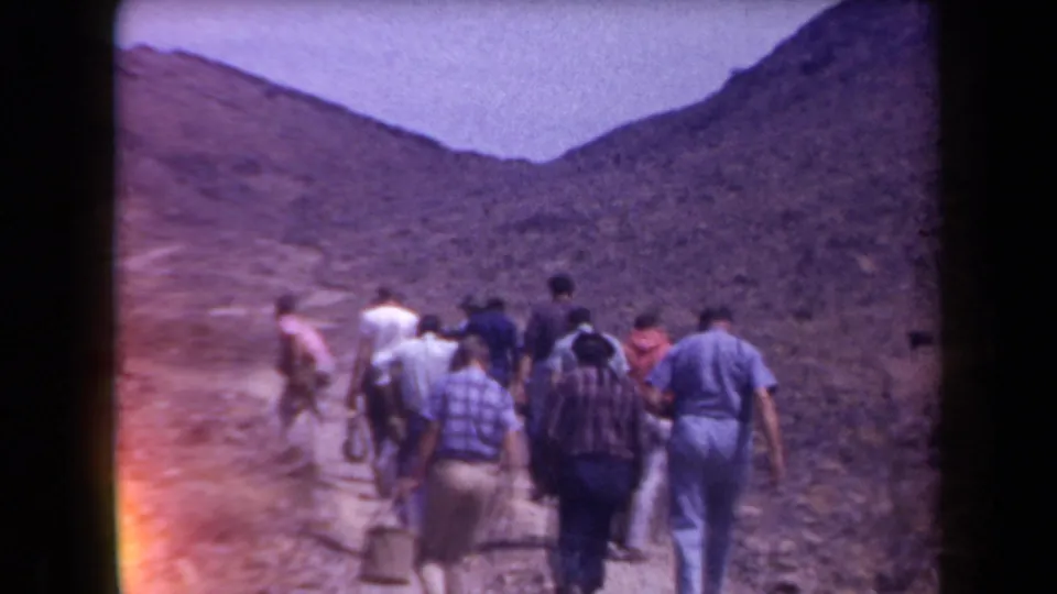 Adult men hike through an Arizona desert valley in 1958, with rocky hills around them and a metal pail visible among the group.