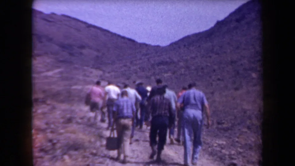 Adult hikers walk away from the camera on an Arizona mountain path, with dry slopes and sparse vegetation around them.