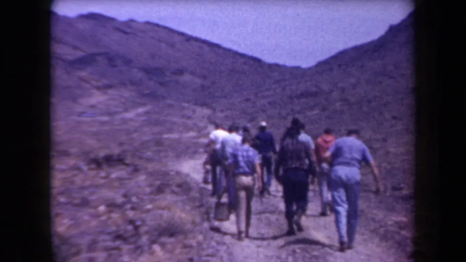 1950s home-movie view of hikers seen from behind as they climb a narrow trail through arid Arizona hills.