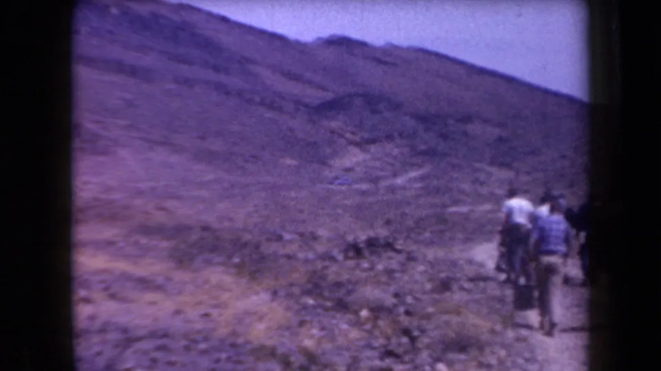 Adult hikers appear near the edge of frame on a rocky Arizona trail, surrounded by wide dry slopes.