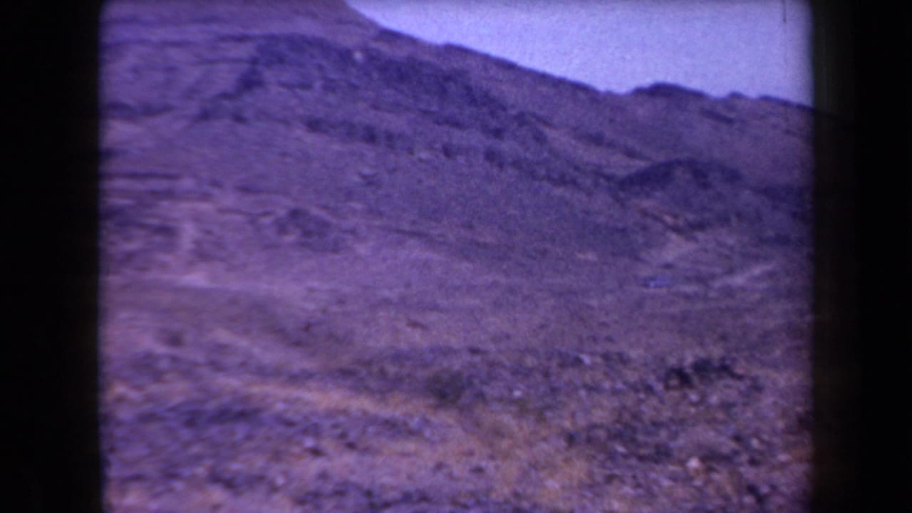 1958 Arizona 8mm frame showing an arid rocky hillside with scrub and a layered mountain ridge under a pale sky.