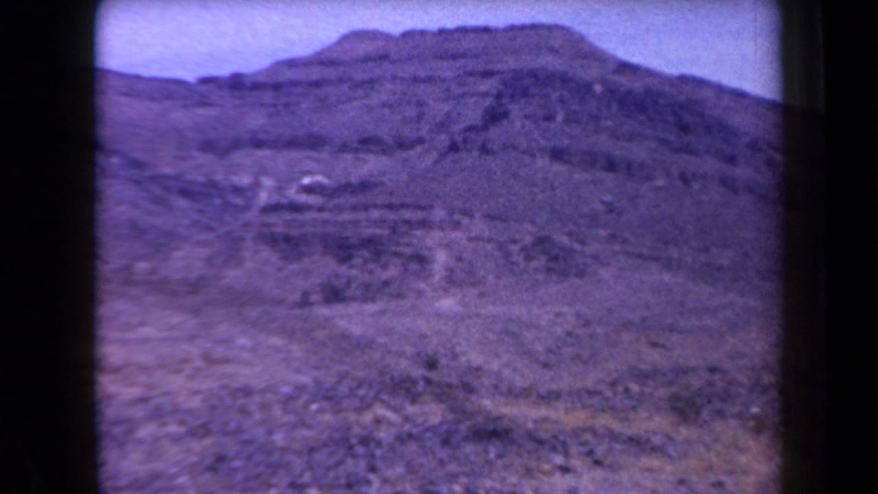 1958 Arizona home-movie frame looking across a rocky desert slope toward layered mountain peaks and sparse scrub.