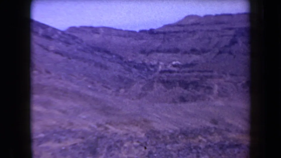 1958 Arizona 8mm frame showing a broad rocky hillside, low desert vegetation, distant ridge, and dark film-frame borders.