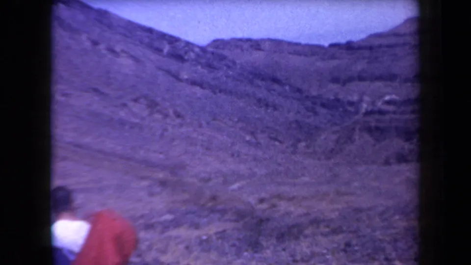 1958 Arizona home-movie frame with a hiker in white and red partly visible before rocky desert slopes and layered ridges.