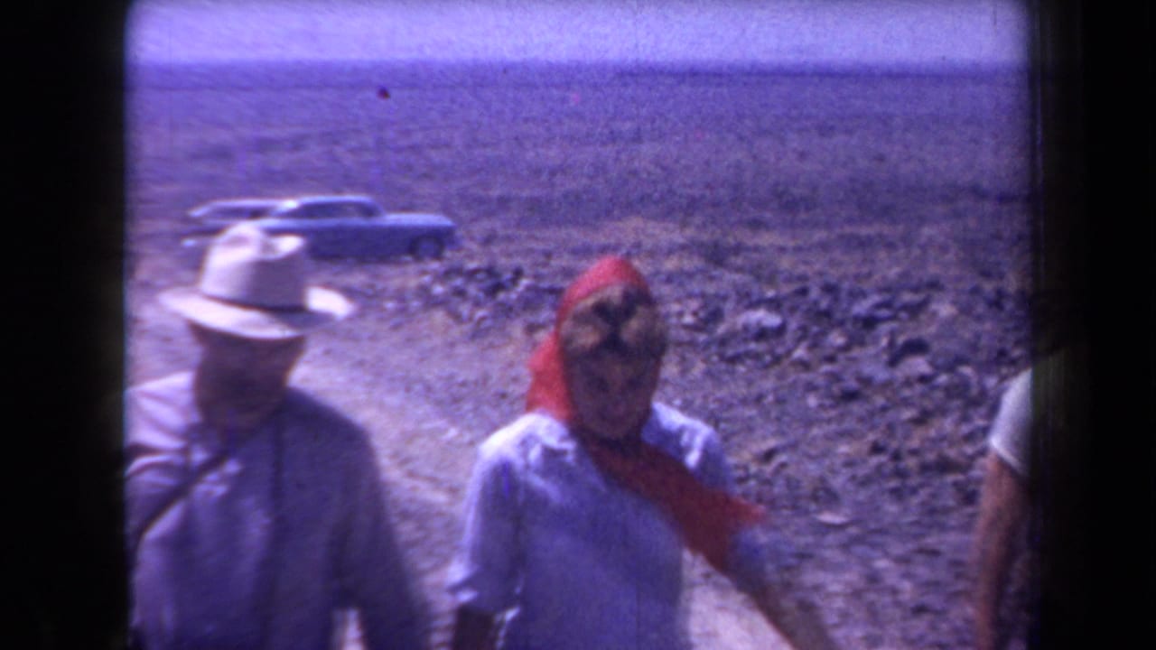 In 1958 Arizona home-movie footage, adults in hats and light clothing walk near a rocky desert road with cars behind them.