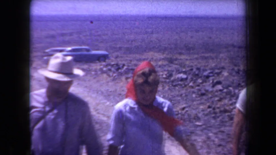 In 1958 Arizona home-movie footage, adults in hats and light clothing walk near a rocky desert road with cars behind them.