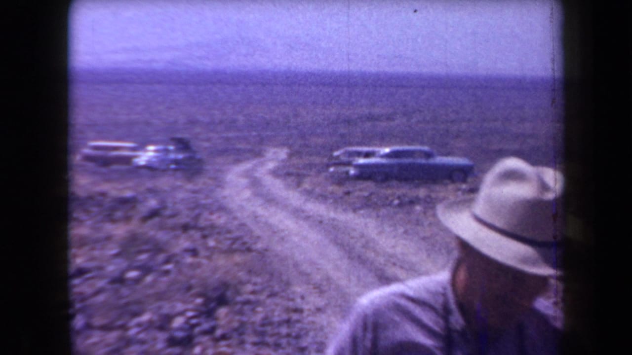 A man in a brimmed hat appears near a winding dirt road in Arizona desert terrain, with late-1950s cars parked behind him.