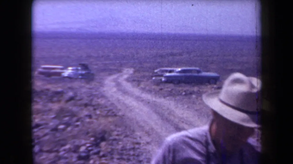 A man in a brimmed hat appears near a winding dirt road in Arizona desert terrain, with late-1950s cars parked behind him.
