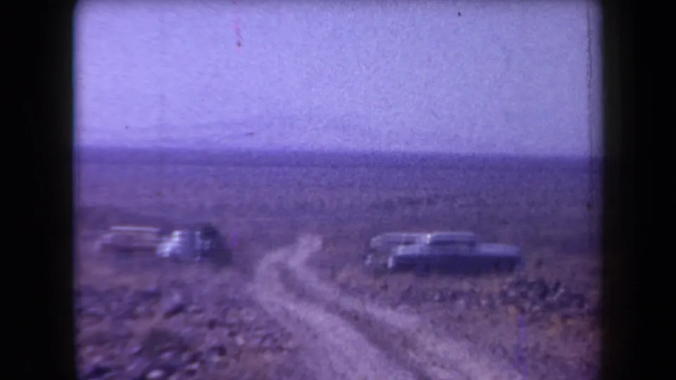 Wide home-movie view of a dusty Arizona desert road with late-1950s cars parked beside the track.