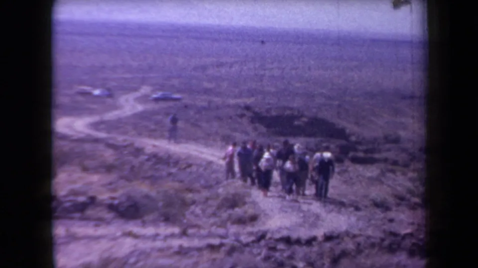 A group of adults walks down a winding dirt road in the Arizona desert, with cars parked farther back.