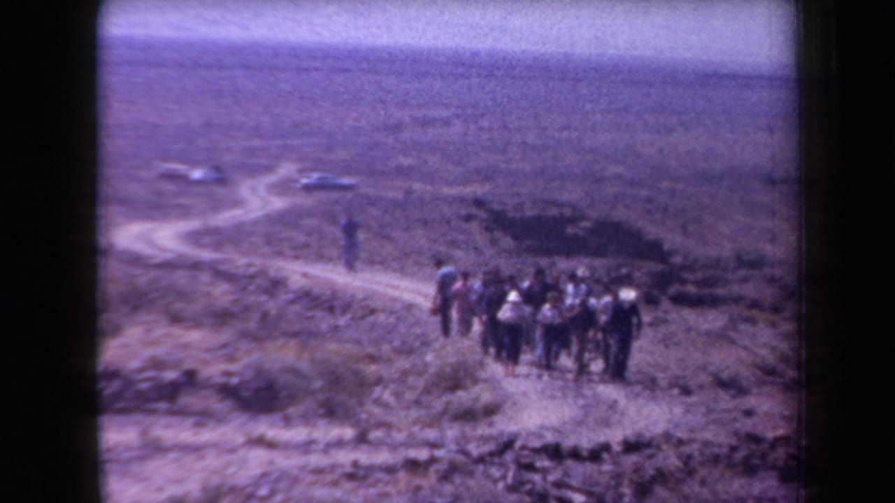 1958 Arizona home-movie frame showing men hiking across dry desert terrain with cars parked in the distance.