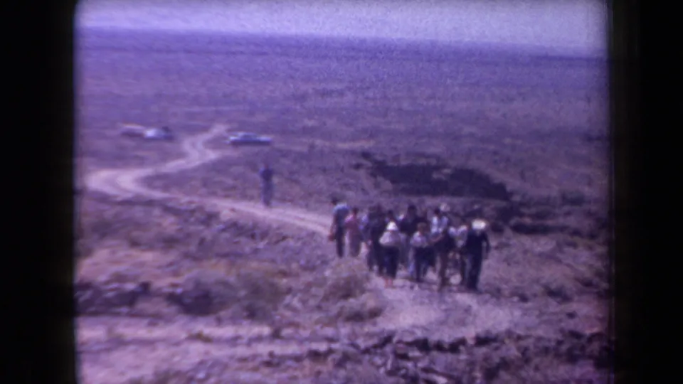 1958 Arizona home-movie frame showing men hiking across dry desert terrain with cars parked in the distance.