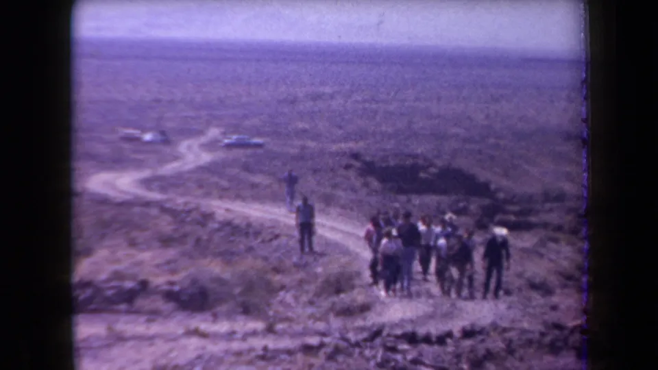 1958 Arizona home-movie frame showing hikers paused on open desert ground with distant cars and sparse vegetation.