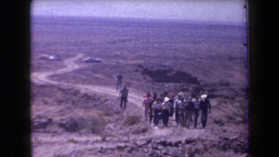 1958 Arizona desert footage frame with men standing in a loose line below a hazy sky and distant road.