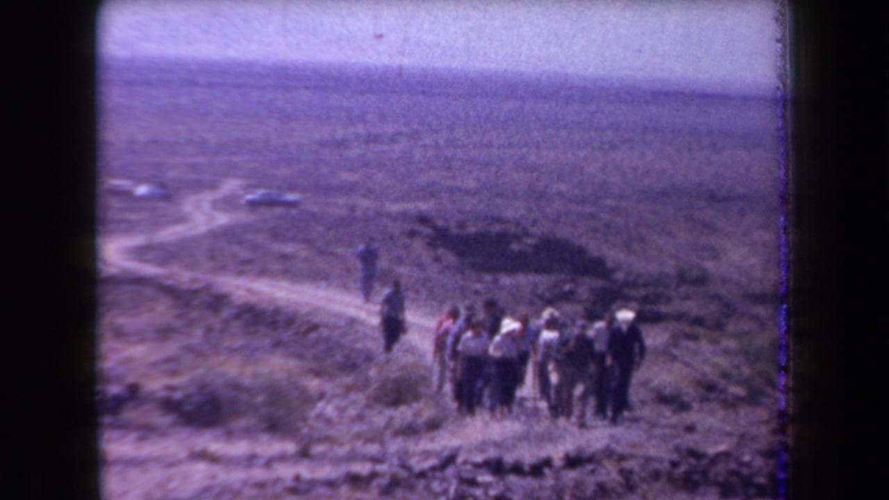 1958 Arizona 8mm frame showing adults on rugged desert terrain, with a dirt road and parked cars beyond them.