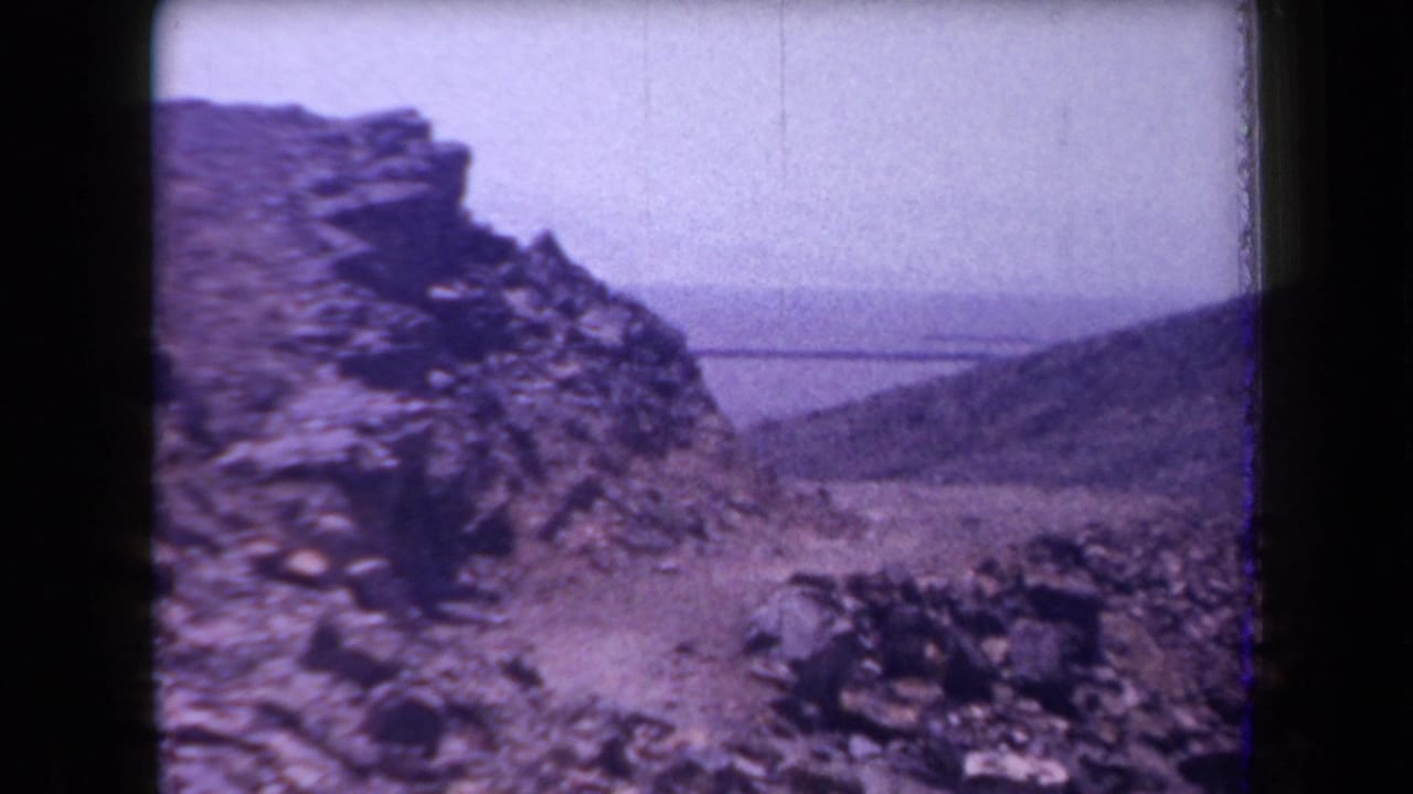 1958 Arizona 8mm view of a rugged stone slope with arid hills and a distant body of water beyond.