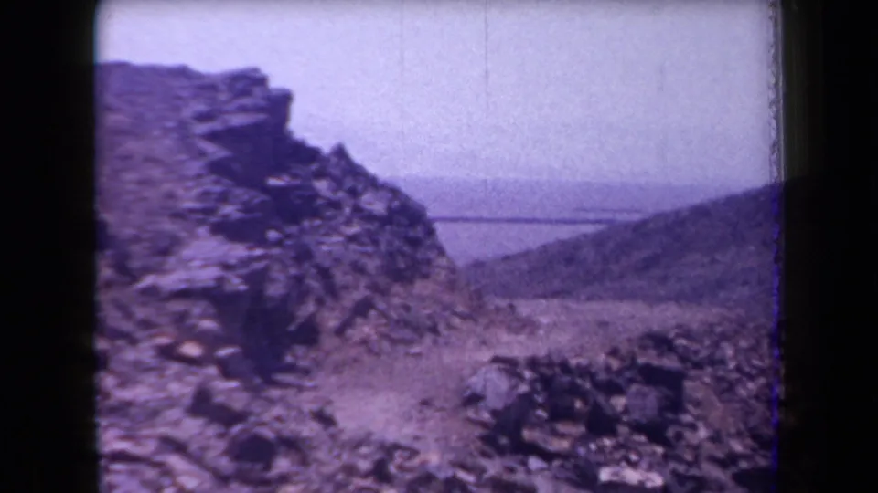 1958 Arizona 8mm view of a rugged stone slope with arid hills and a distant body of water beyond.