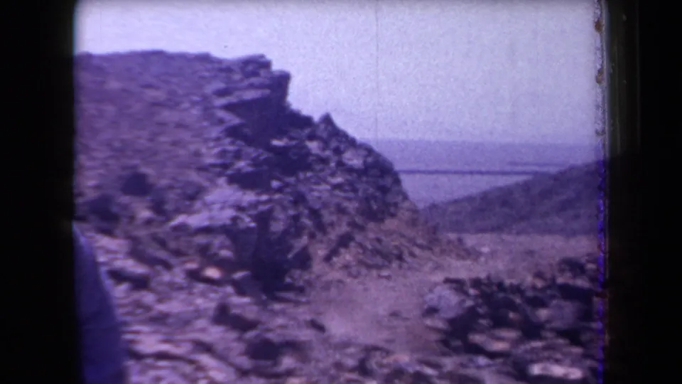 1958 Arizona 8mm frame with a partial figure near a stone-covered slope and a distant body of water.