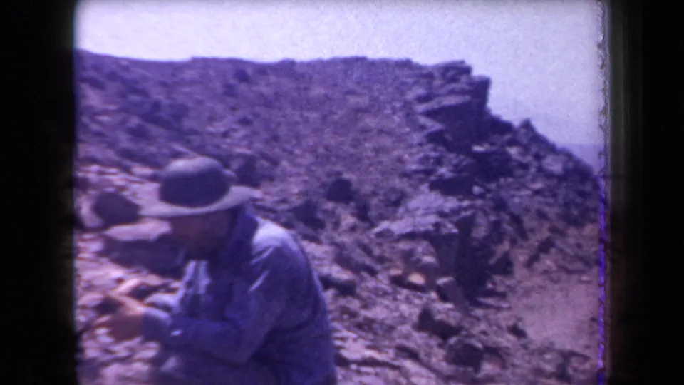 1958 Arizona 8mm frame showing a man in a hat crouched close to stones on rugged desert ground.