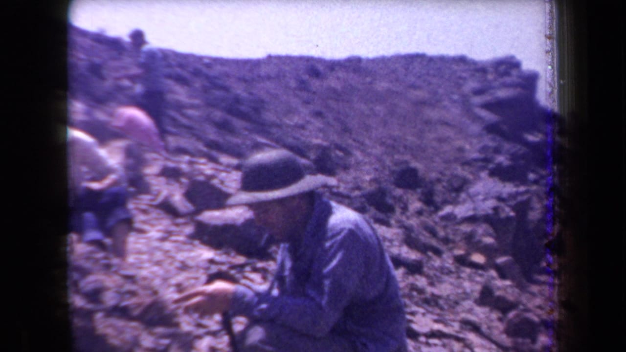 1958 Arizona 8mm frame showing adults in hats and light clothing working or examining ground on a rocky arid slope.