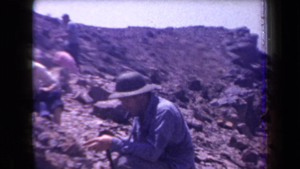 1958 Arizona 8mm frame showing adults in hats and light clothing working or examining ground on a rocky arid slope.