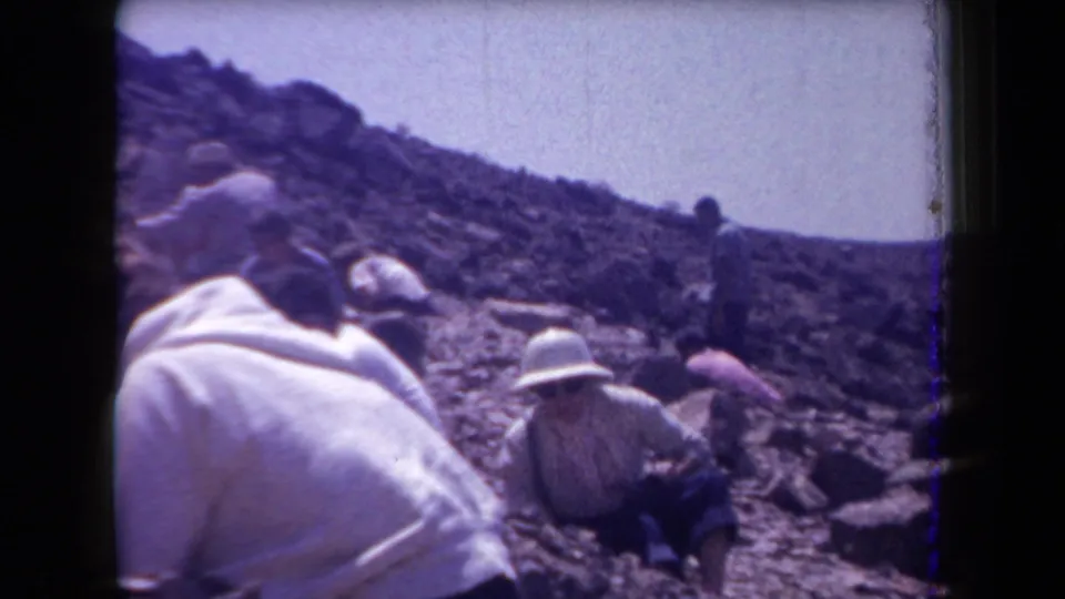 1958 Arizona home-movie view of adults scattered across an arid rocky slope, with figures crouching and leaning near the ground.