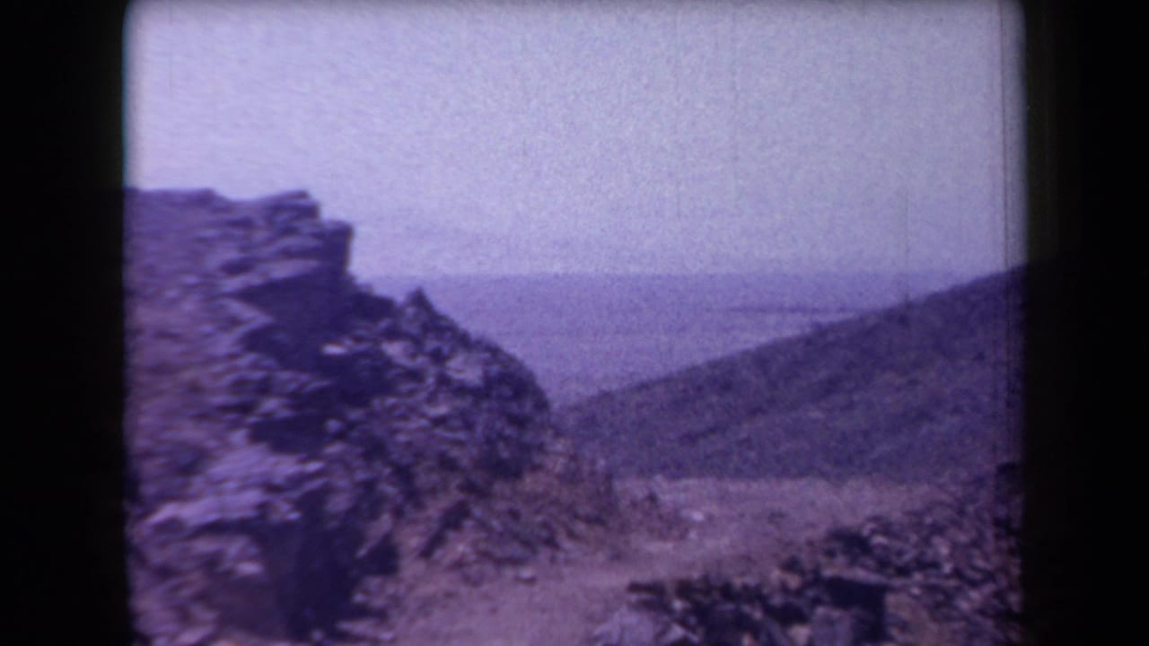 1958 Arizona 8mm frame showing a rocky desert slope, distant landscape, and rough stone formations under bright sun.