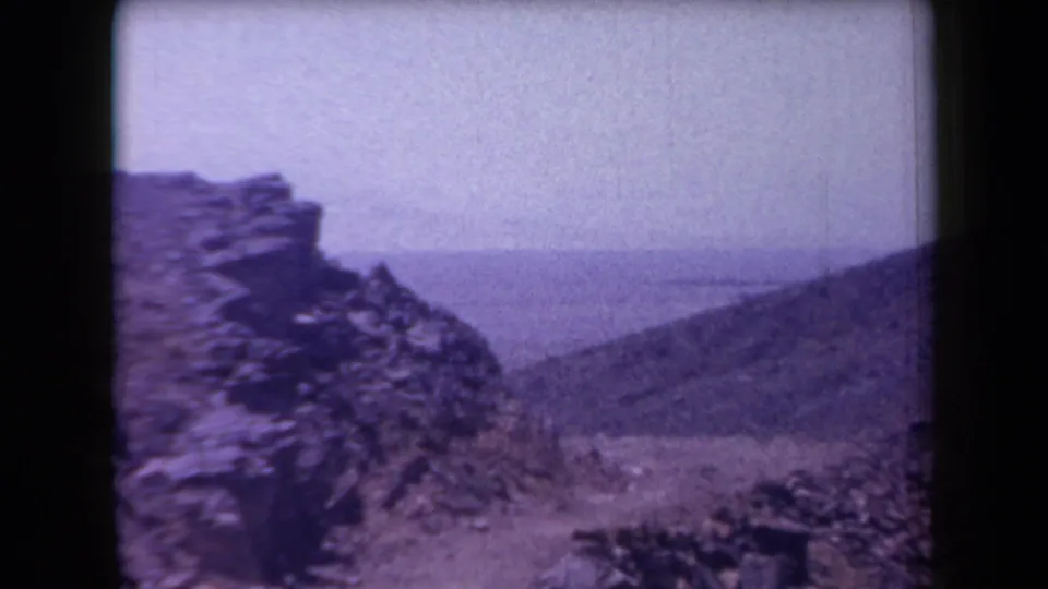 1958 Arizona 8mm frame showing a rocky desert slope, distant landscape, and rough stone formations under bright sun.