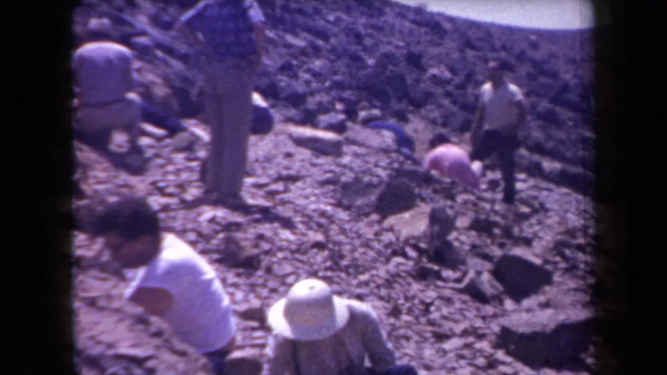 A small group in period outdoor clothing and hats moves through a rocky Arizona dig area in 1958 8mm footage.