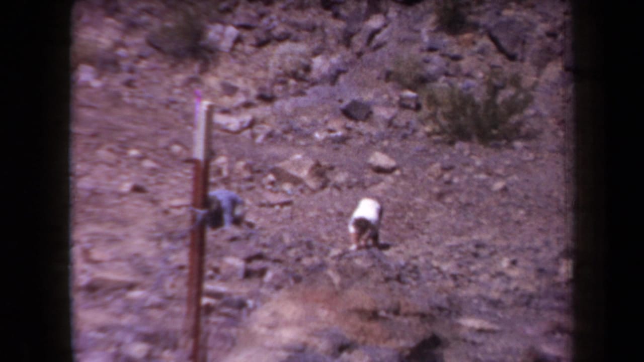 1958 Arizona 8mm frame showing distant people bent toward the ground in a barren, rock-strewn desert area near a vertical pole.