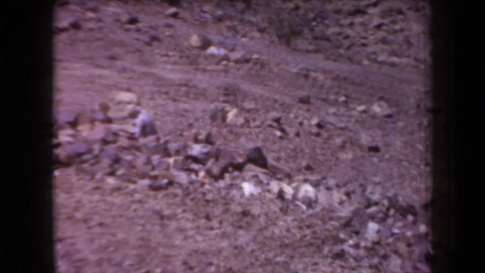 1958 Arizona 8mm frame showing a distant worker bent near a rocky wash, surrounded by dry ground and scattered stones.