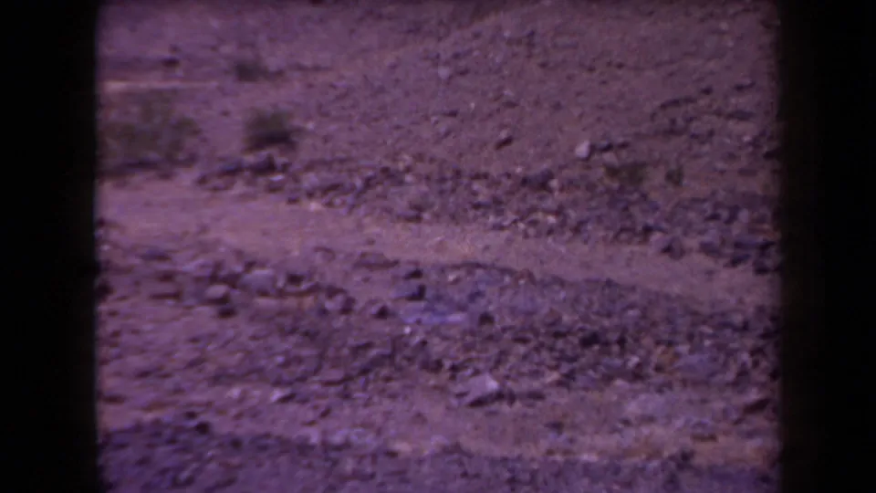 1958 Arizona 8mm view of dry rocky dirt terrain with sparse scrub, magenta fading, and visible film gate edges.