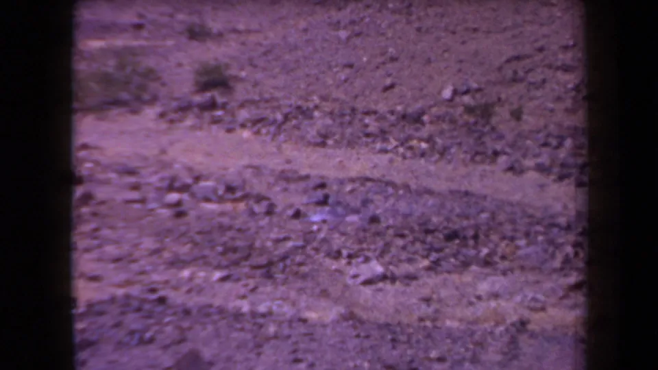 1958 Arizona 8mm frame showing rocky desert terrain, low scrub, and a pale dirt wash with purple color fading and dark gate edges.