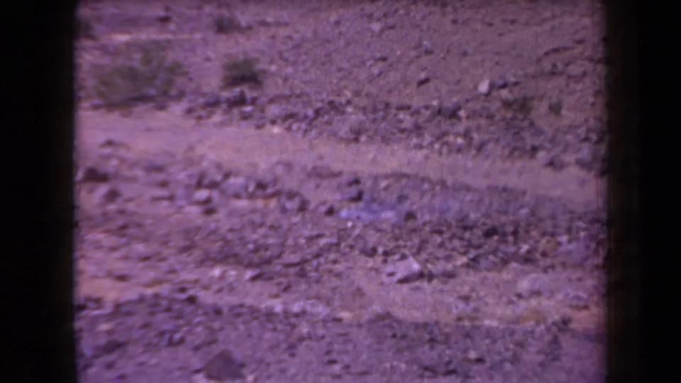 1958 Arizona 8mm still showing a rocky desert surface, sparse vegetation, soft focus, and dark film borders around the scanned frame.