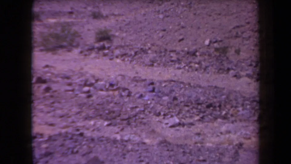 1958 Arizona 8mm frame of a dry dirt field with rocks, scrub, film grain, and magenta-purple color fading.