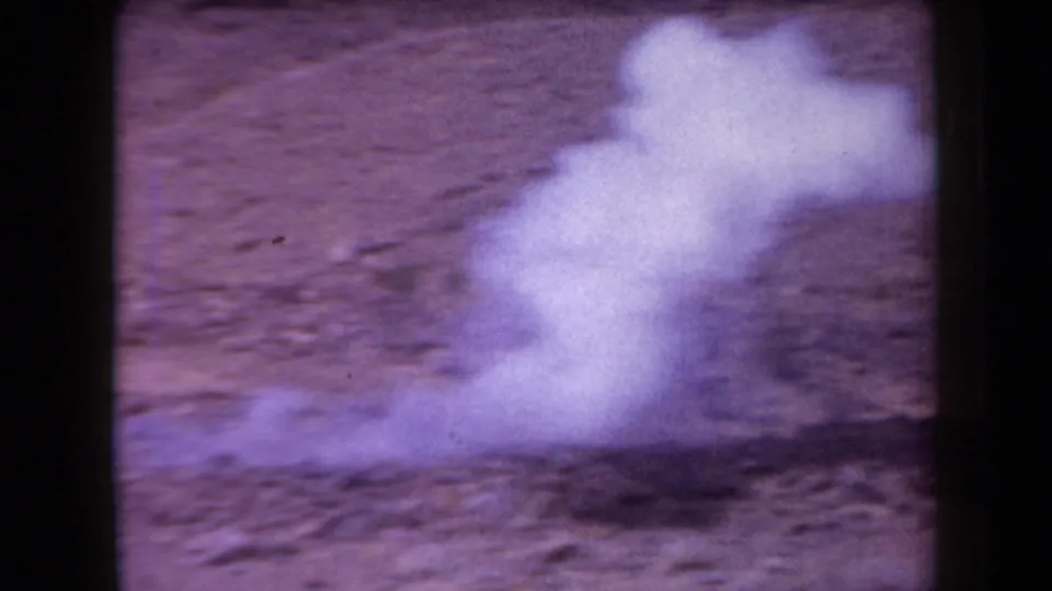 1958 Arizona 8mm frame showing a small white smoke plume spreading low across rocky dirt in an arid field.