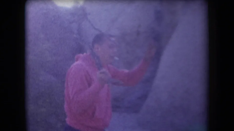 In soft 8mm color, a person in a pink top appears to hold a stick while standing close to a rocky desert surface in Arizona.