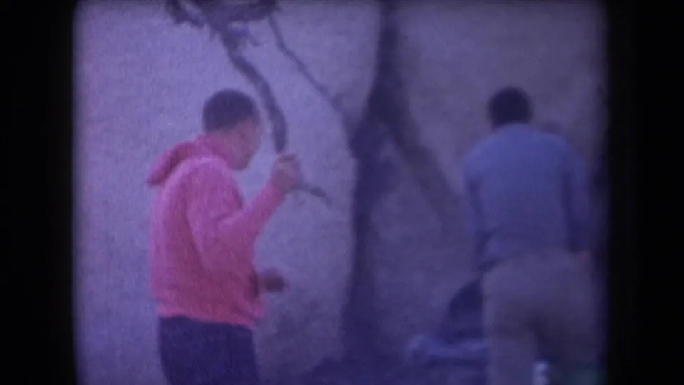 Two people, one in pink and one in a light blue shirt, move beside large rocks in a hazy 1958 Arizona desert home-movie frame.