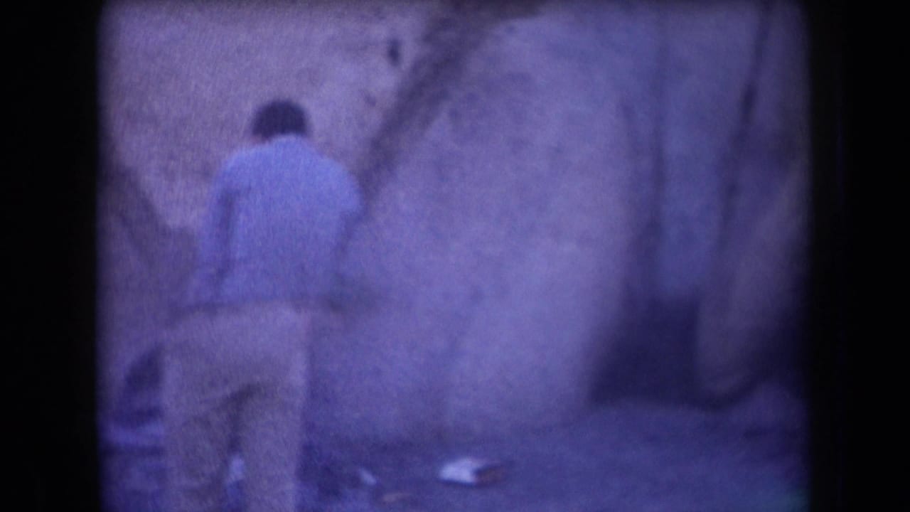 In 1958 Arizona home-movie footage, a man in a light blue shirt stands near a rough rock wall with small items on the ground nearby.