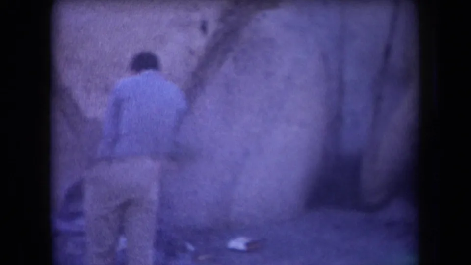 In 1958 Arizona home-movie footage, a man in a light blue shirt stands near a rough rock wall with small items on the ground nearby.