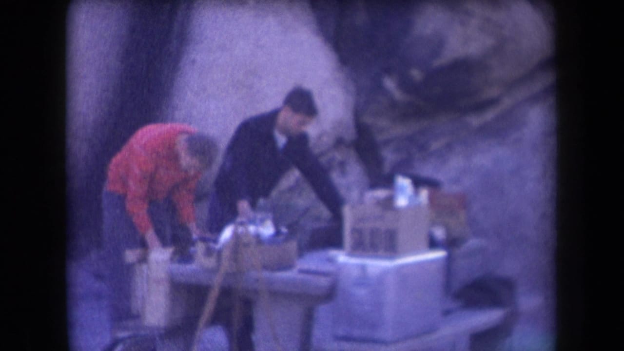 Two adult men in 1958 Arizona appear to sort boxes and containers on an outdoor table or work surface beside a rock wall.