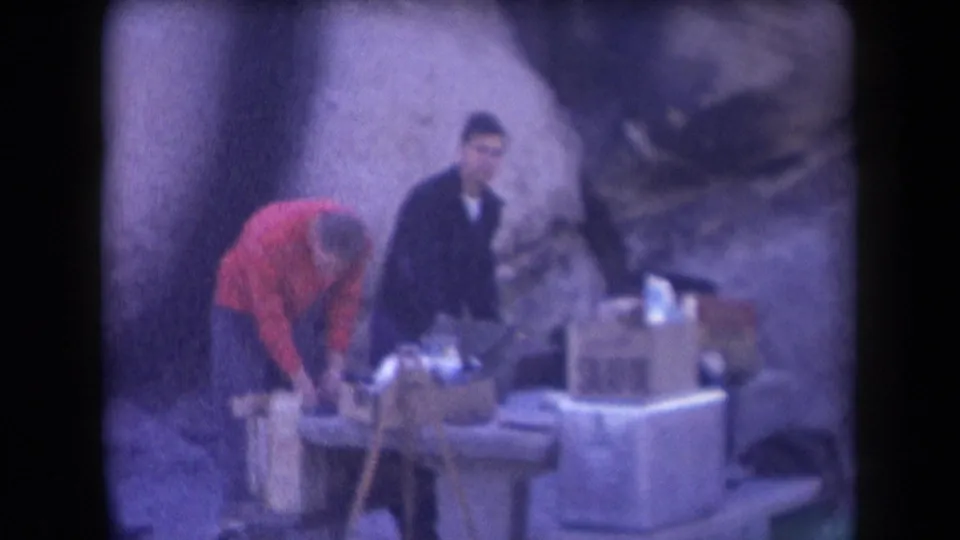 A man in a red sweater bends over supplies on an outdoor table while another man stands nearby in grainy 1958 Arizona 8mm footage.