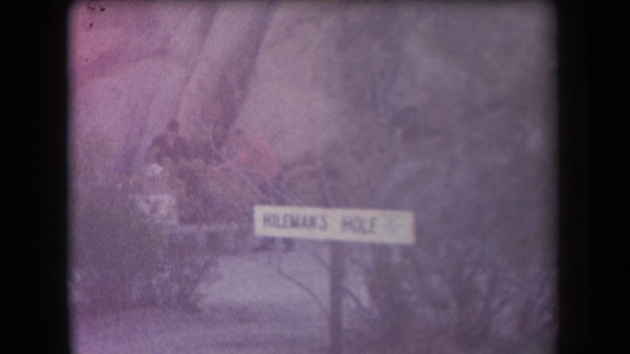 1958 Arizona home-movie frame showing a Hileman's Hole sign amid brush, with distant visitors partly visible behind it.