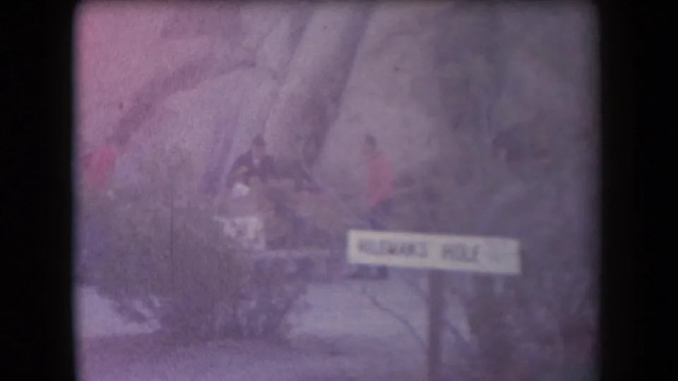 Faded 1958 Arizona 8mm view of the Hileman's Hole sign with blurred visitors partly hidden by brush.