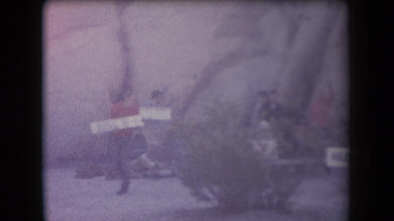 Soft, purple-tinted 1958 Arizona frame with people gathered near a rocky site and a partially visible sign.