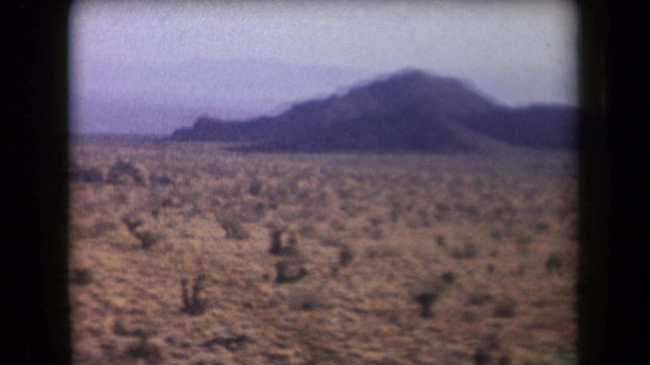 1958 Arizona 8mm frame showing a rocky desert plain with sparse shrubs and bluish hills in the distance.