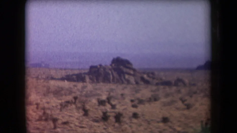 1958 Arizona 8mm frame showing scrubby desert foreground, a rocky formation, and hazy hills on the horizon.