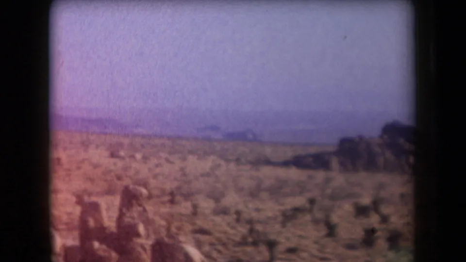 1958 Arizona 8mm frame with sparse desert plants, hazy bluish hills, and a dark rock outcrop near the right edge.